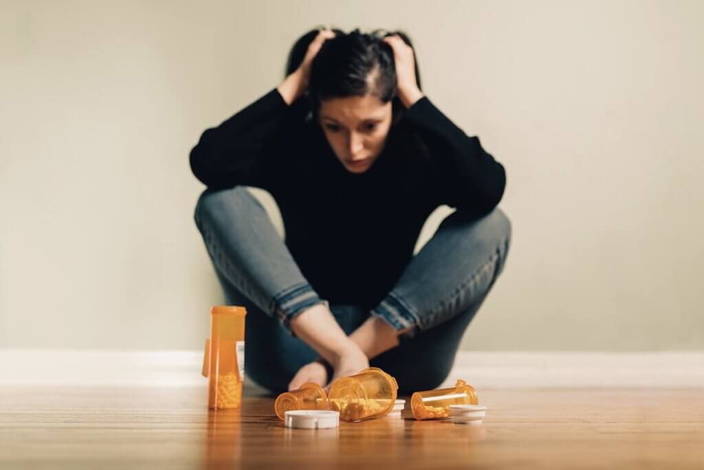 girl sitting on the floor with an open bottle of pills in front of her is holding her head in pain, she is in desperate need of an inpatient heroin rehab center for help.